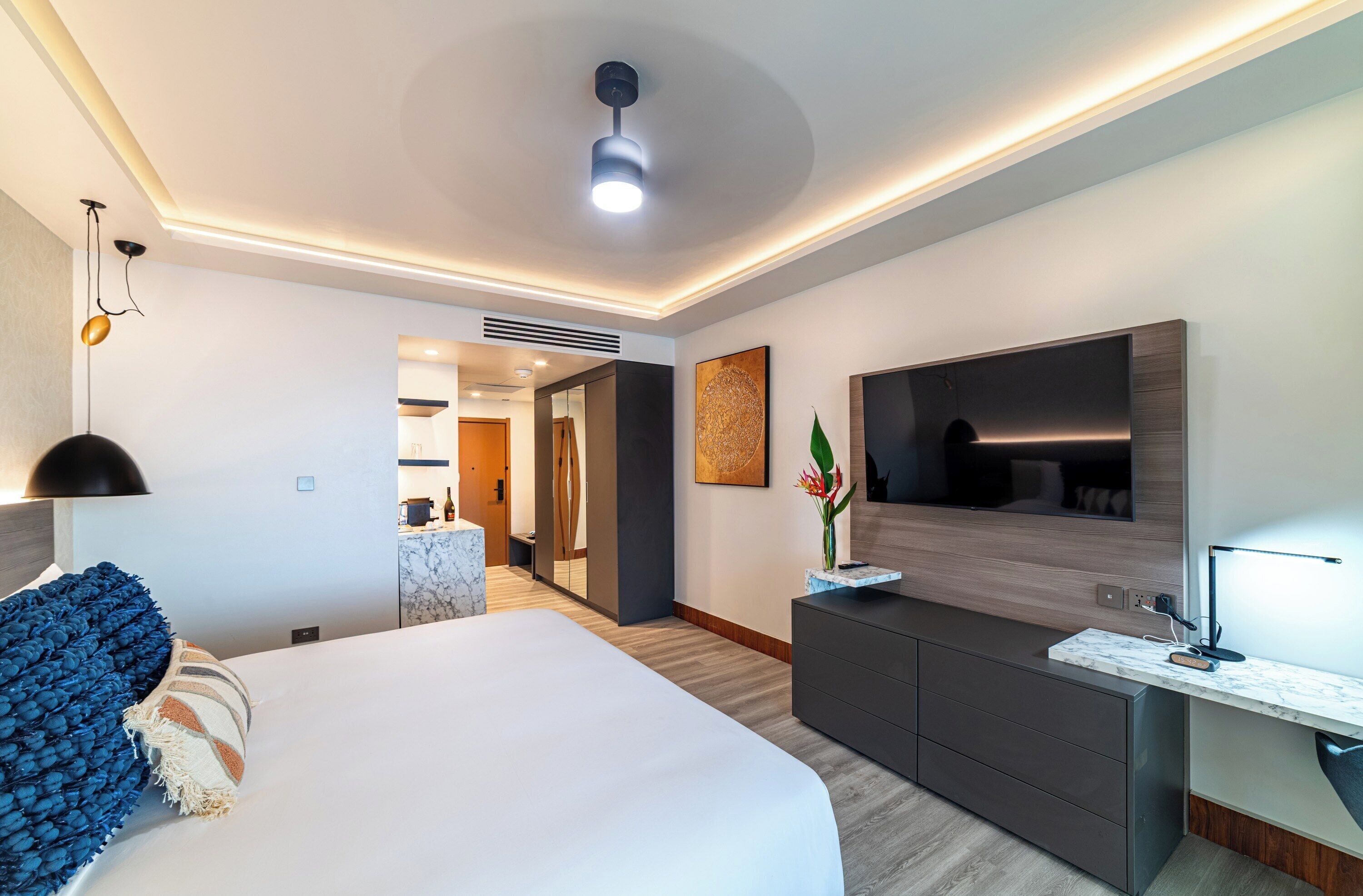 A modern hotel room with a large bed, a desk with a flat-screen TV, a ceiling fan, and a hallway leading to a bathroom.