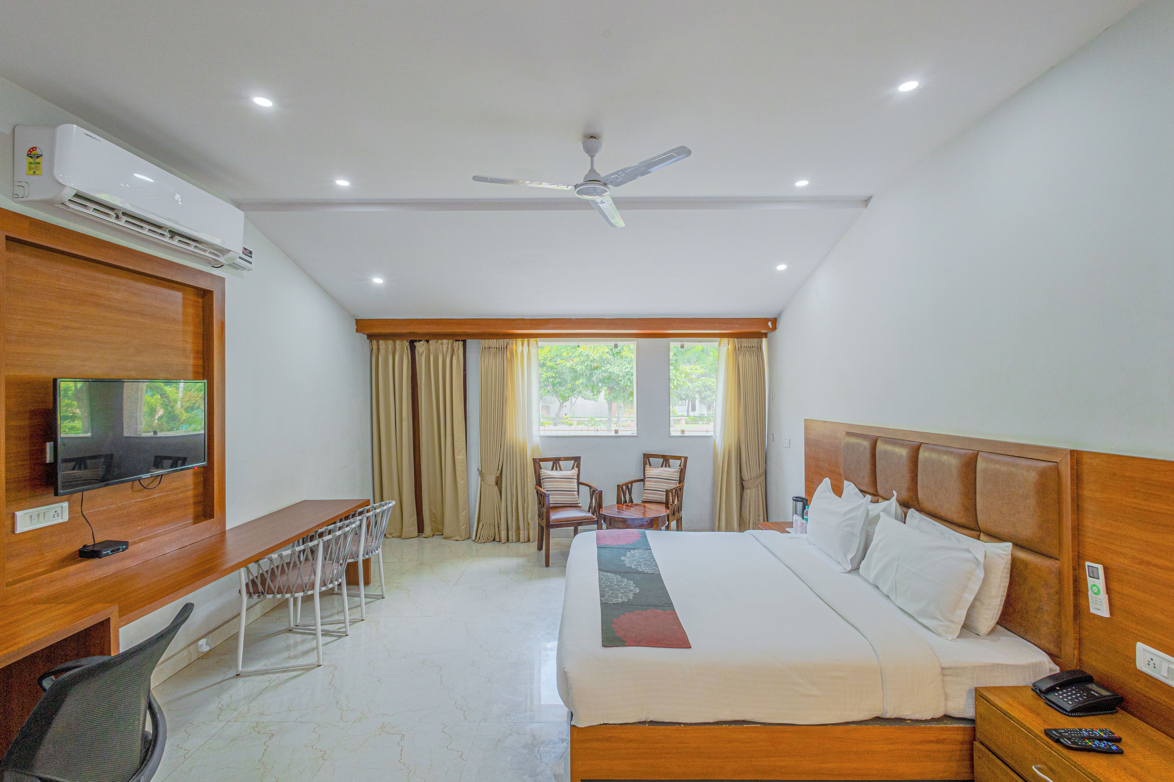 A hotel room with a bed, desk, chairs, TV, and an air conditioning unit.