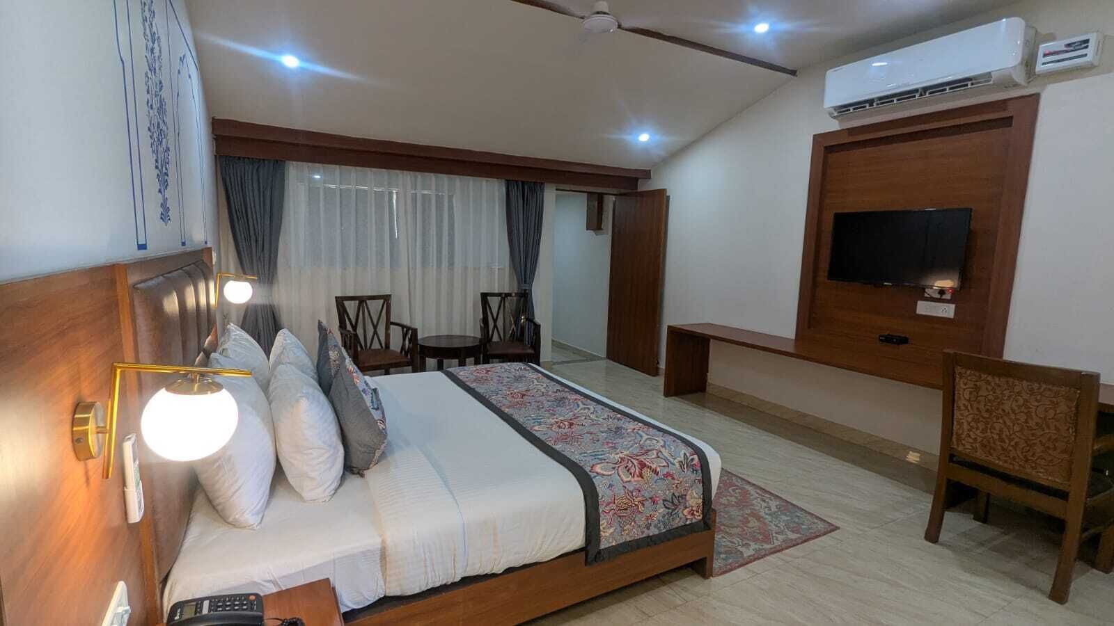 A hotel room with a bed, bedside lamp, TV, air conditioning unit, and a small seating area.