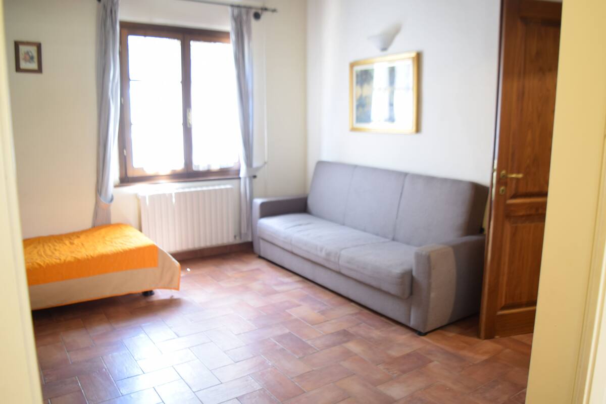 A bedroom with a bed, a grey sofa, a window with curtains, and a radiator.