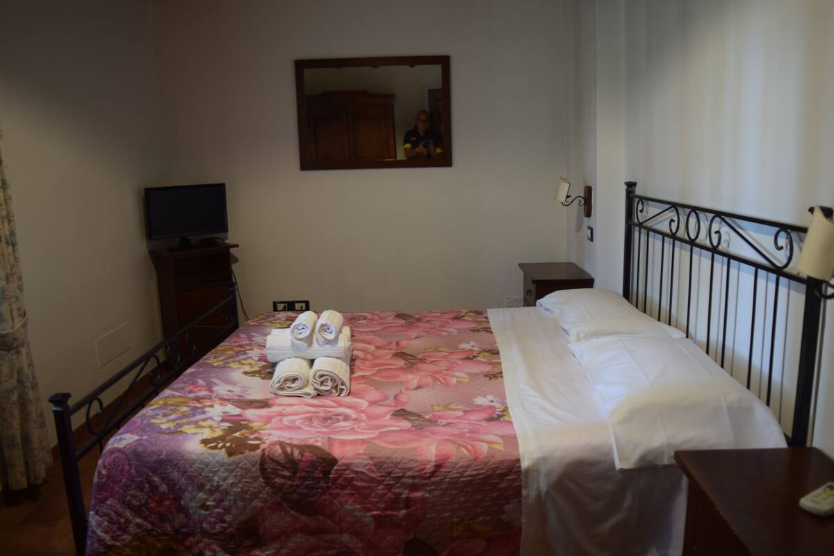 A hotel room with a bed, a TV, a mirror, and a bedside table.