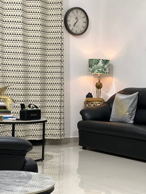 A modern living room with a black leather sofa, a round coffee table, a patterned curtain, a wall clock, a lamp with a leaf design, and a small table with a speaker.