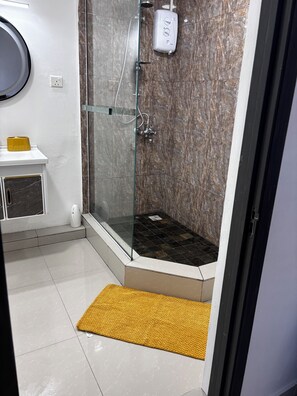 Ein Badezimmer mit einer in Glas abgeschirmten Dusche, einer marmorverkleideten Wand, einer gelben Matte und einem weißen Waschbecken mit gelbem Seifenspender.