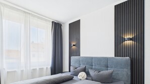 Una habitación de hotel moderna con una cama grande, ropa de cama gris, cabecera con líneas verticales y una lámpara fijada a la pared.