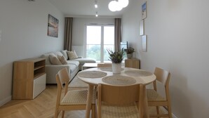 Uma sala de estar moderna com sofá, mesa de jantar com cadeiras, uma planta em vaso e uma grande janela com cortinas.