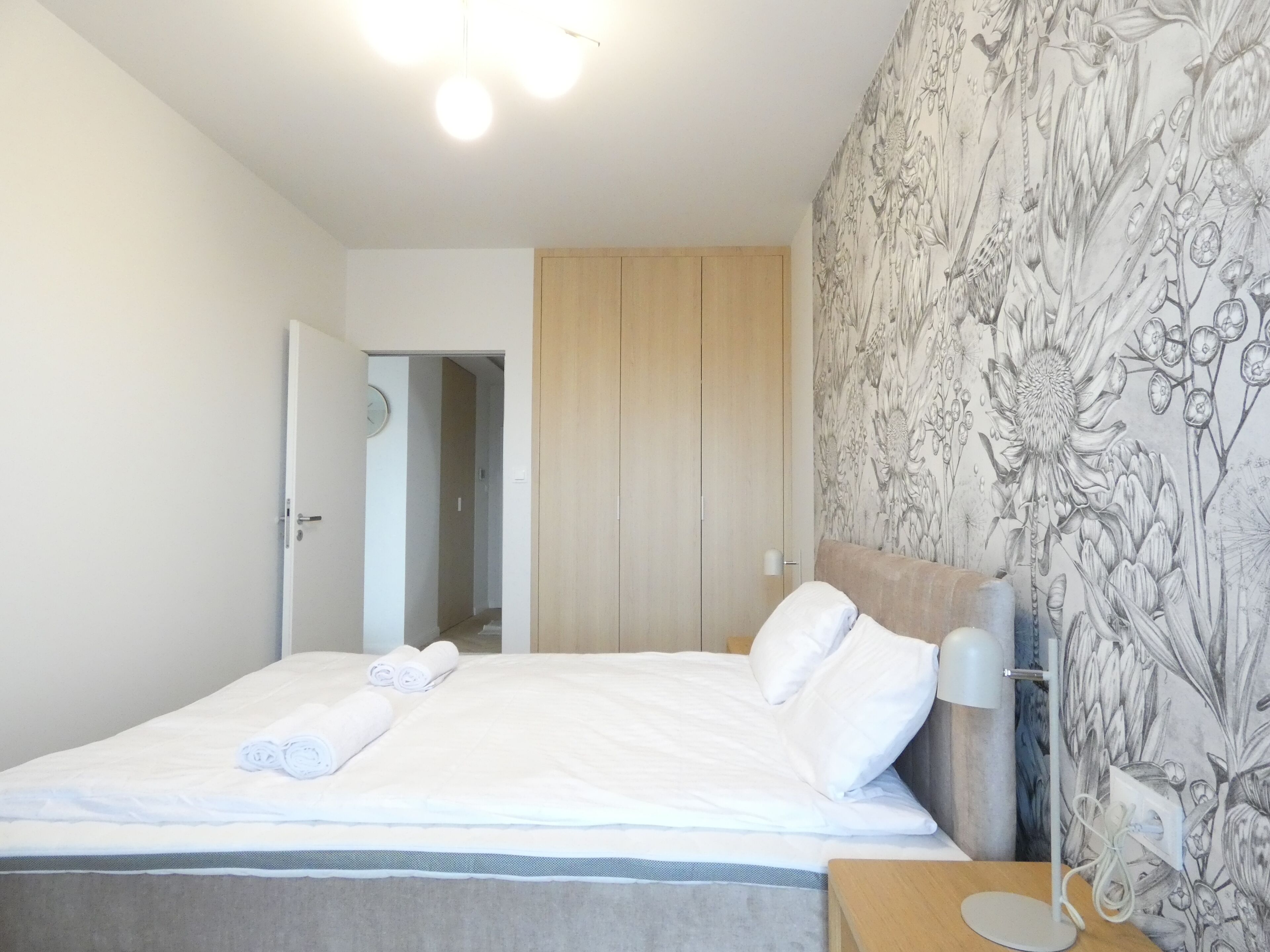 A hotel room with a bed, a nightstand, a lamp, a wooden wardrobe, and a floral wallpaper.
