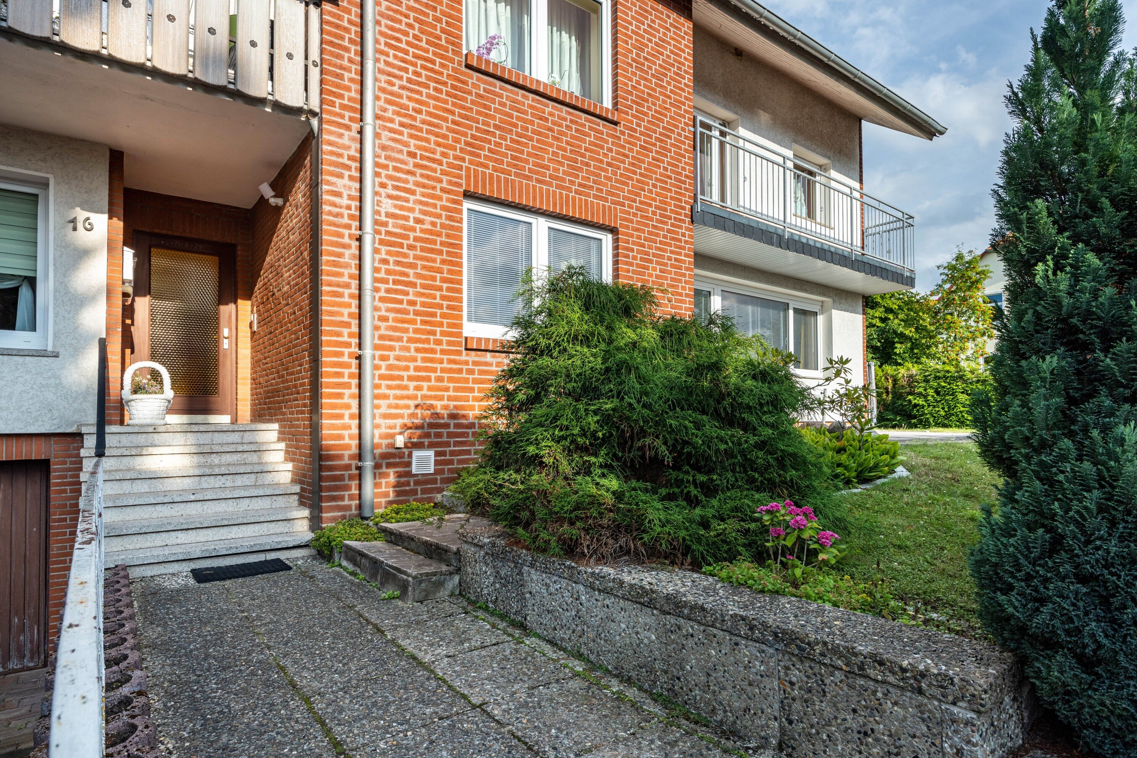 Un immeuble résidentiel de deux étages, doté d’une façade en briques, d’un balcon à l’étage supérieur et d’un petit jardin devant, agrémenté d’arbustes et de fleurs.