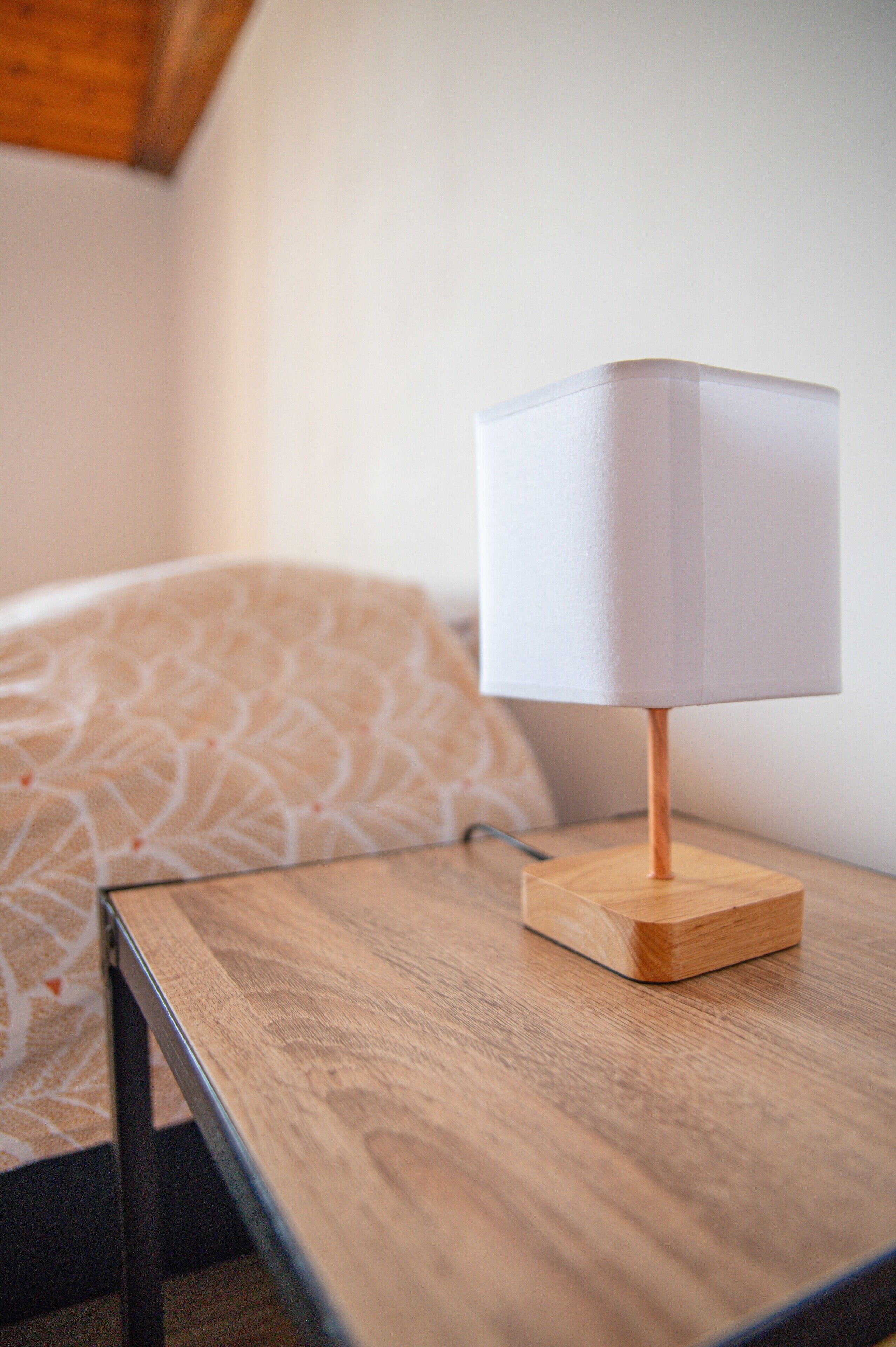 Une table de chevet en bois avec une lampe blanche, une couvre-lit à motifs et une tête de lit en bois, dans une chambre d’hôtel.