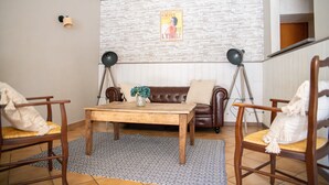 Ein gemütliches Wohnzimmer mit einer braunen Ledersofa, einem Holztisch und zwei Holzstühlen mit Kissen. Auf dem Boden liegt ein gemustertes Teppich, und an der Wand ist ein Poster zu sehen.