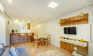 A modern living room with a sofa, a dining area with chairs, a TV mounted on the wall, and a wooden cabinet with decorative items. - Apart w/ 2 Bedrooms and Fireplace - Canela Epe0304 (Canela)