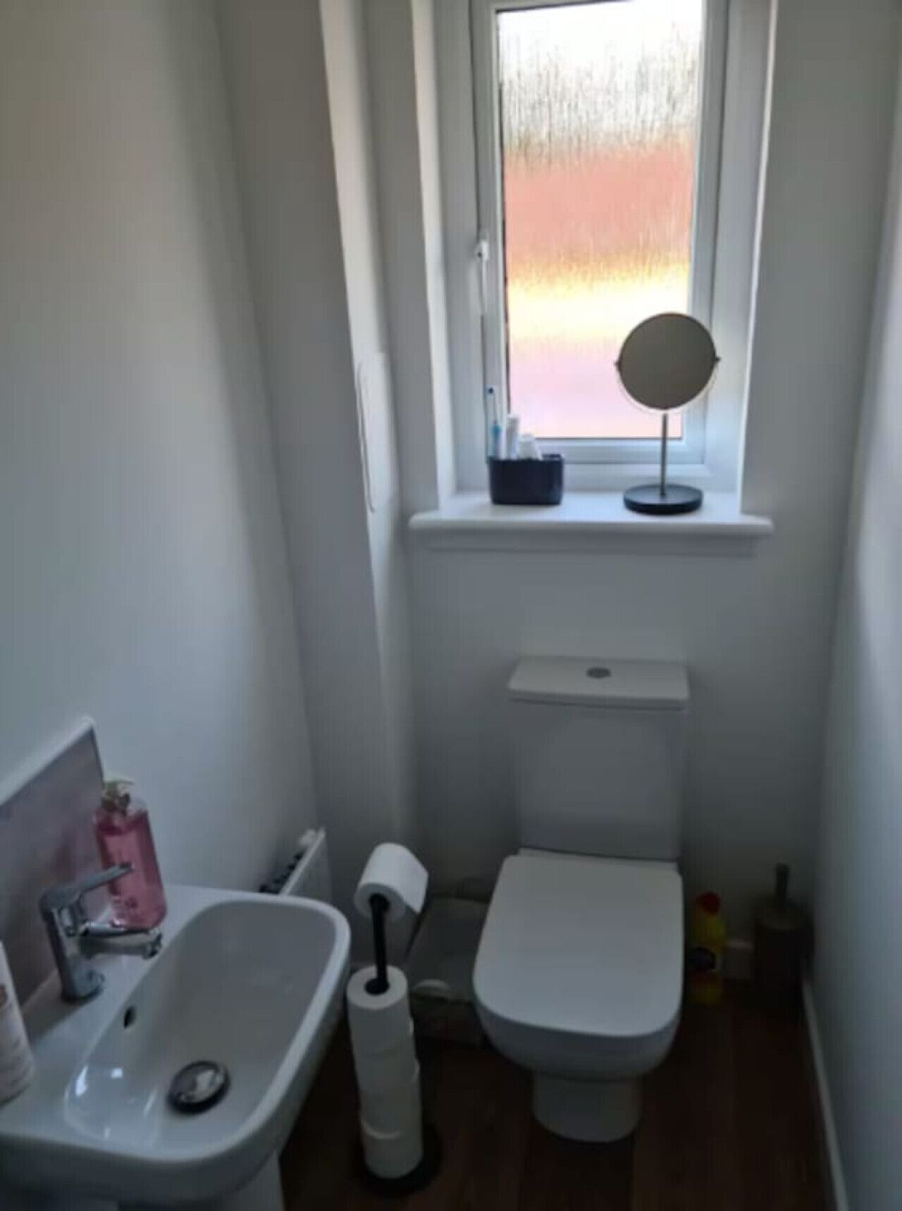 A small, compact bathroom with a window, a sink, a toilet, and a mirror on the windowsill.