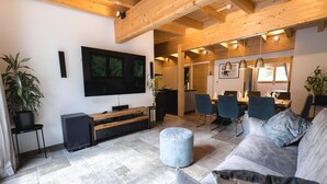 A modern living room with a large flat-screen TV, wooden beams on the ceiling, a grey sofa, and a dining area with chairs and a table. - Penthouse at 1900 Meters Above sea Level (Soelden)