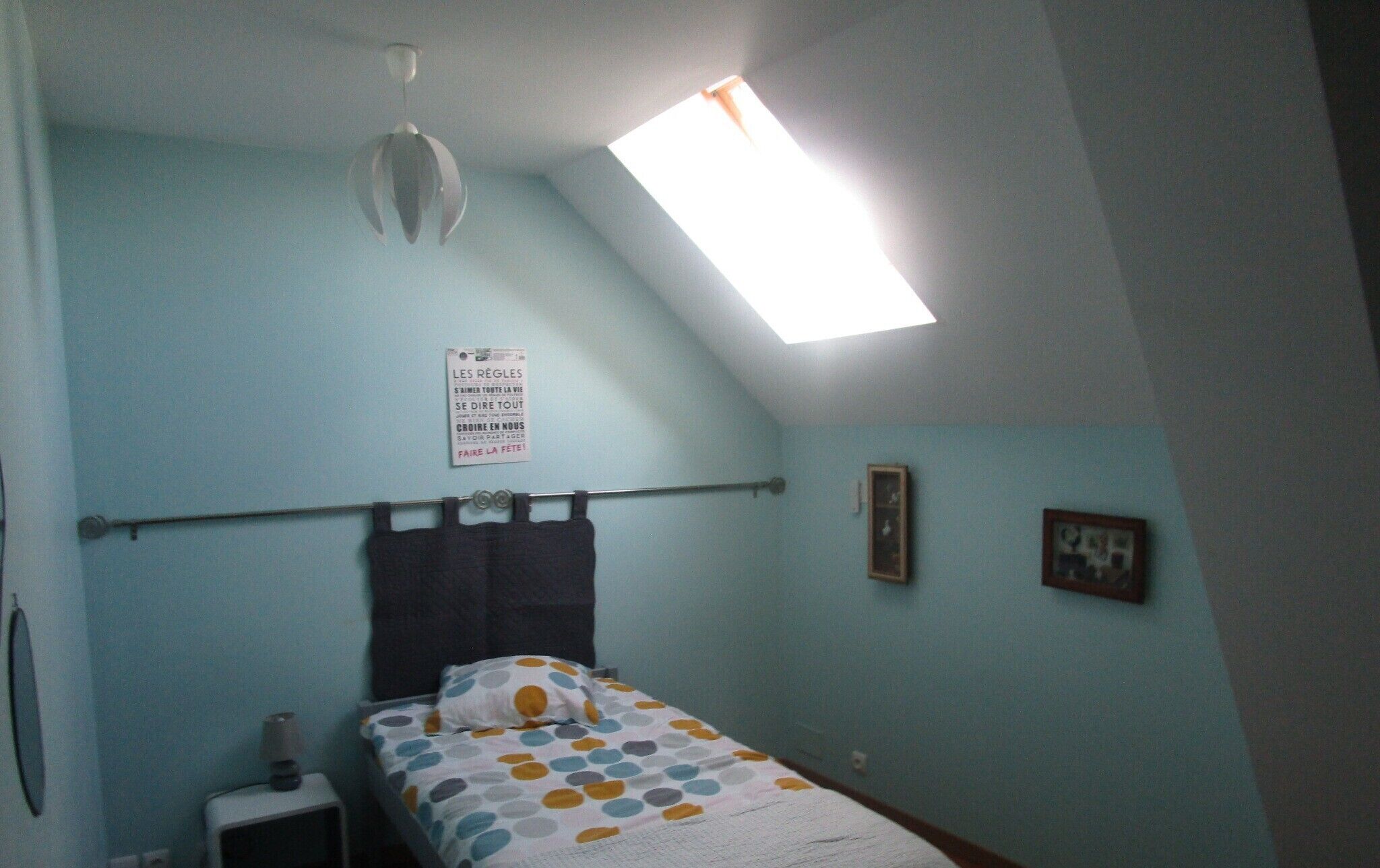 Une chambre à coucher comprenant un lit, un ventilateur de plafond, un puits de lumière, un panneau fixé au mur et une petite table de chevet.