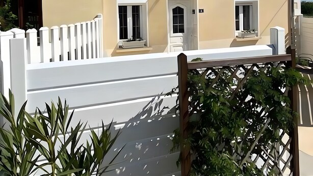 Un bâtiment à deux étages, doté d’une façade beige, d’un toit recouvert de tuiles brunes et d’une clôture blanche en bois.