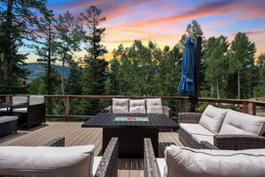 Terraza al aire libre con mobiliario de mimbre, una mesa con cubierta de vidrio y vistas a un área boscosa al atardecer.