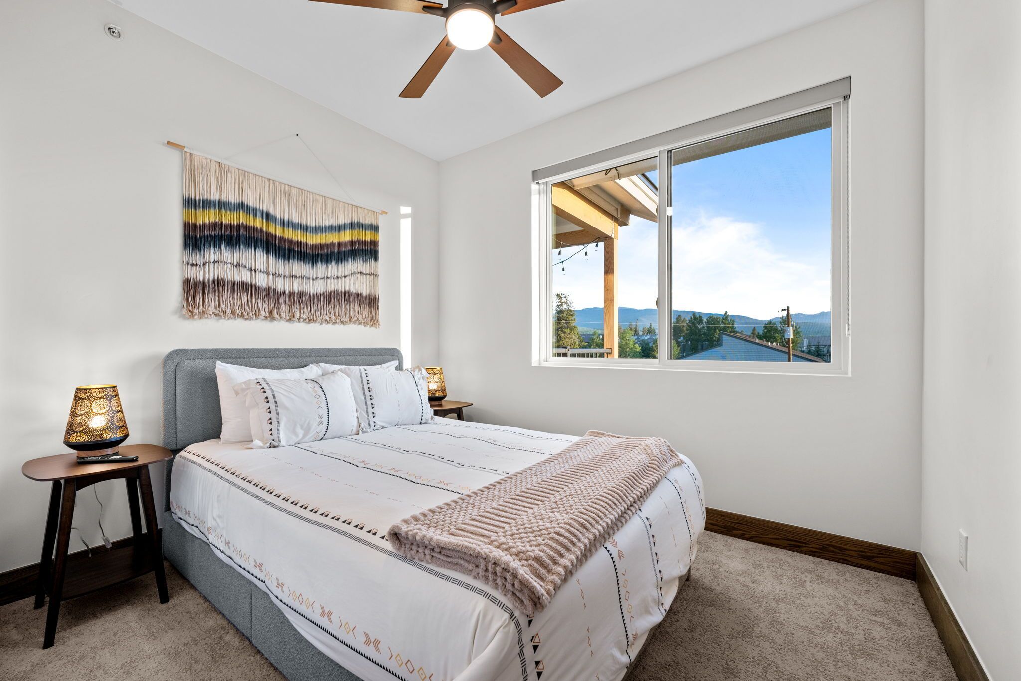 A bedroom with a large bed, a ceiling fan, a wall-mounted tapestry, a bedside lamp, and a window with a view of a landscape.