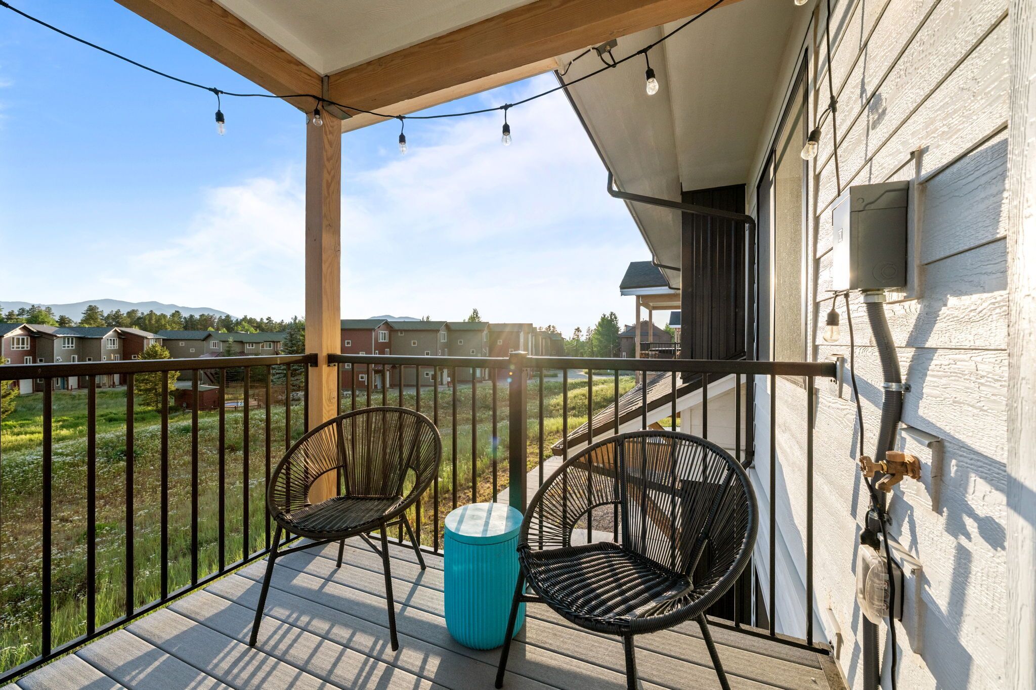 Un balcon avec deux fauteuils en osier et une poubelle bleue, donnant sur un quartier résidentiel avec des maisons et un champ d’herbe.