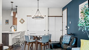 Eine moderne Küche mit weißen Schränken, einem runden Esstisch und Barhocker. Über dem Tisch hängt eine Chandelier und rechts steht ein Blumentopf.
