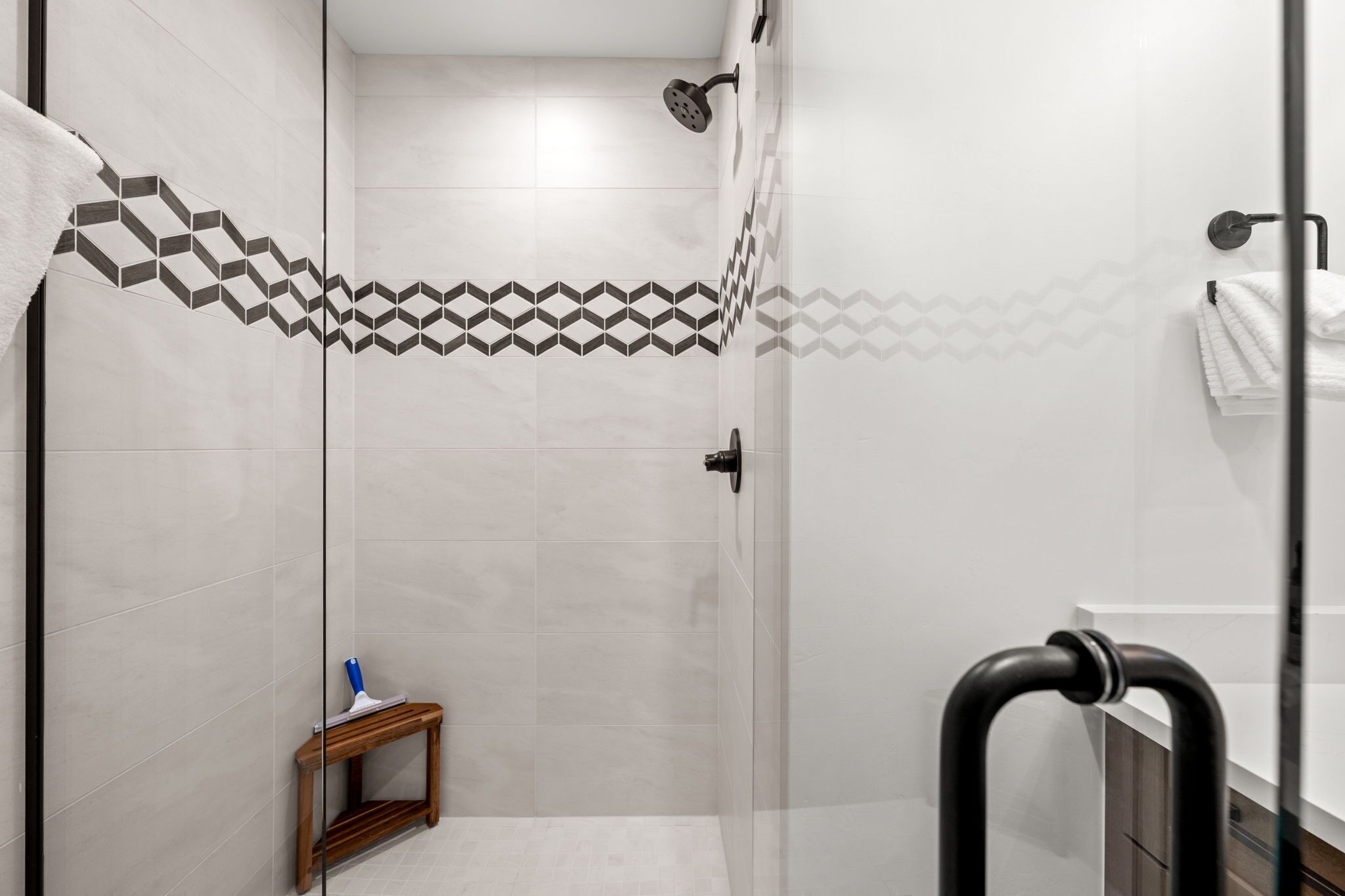 Un espace douche moderne avec des motifs géométriques sur les carreaux, un petit tabouret en bois et une paroi de douche en verre.