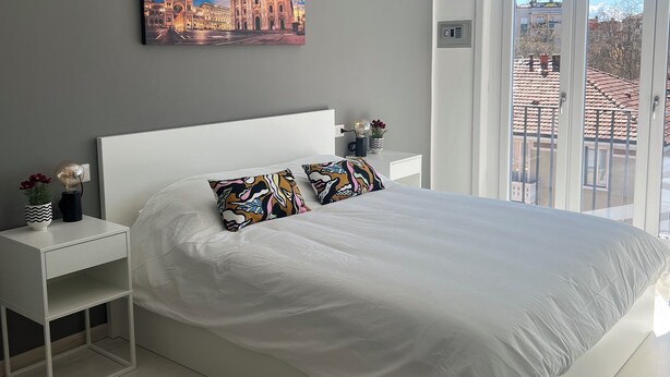 Ein modernes Schlafzimmer mit einem großen Bett, einem Nachttisch, einem Gemälde einer Kathedrale und Blick durch eine Glastür.