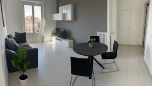 Ein modernes Wohnzimmer mit einem grauen Sofa, einem runden Esstisch mit Stühlen, einem Fernseher auf einem weißen Schrank und einem Blumentopf.