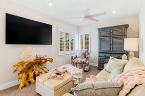 A living room with a flat-screen TV, a ceiling fan, a wooden coffee table, a patterned armchair, a sofa, and a dark wooden cabinet.