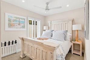 A bedroom with a bed, a ceiling fan, a window with shutters, and a painting on the wall.