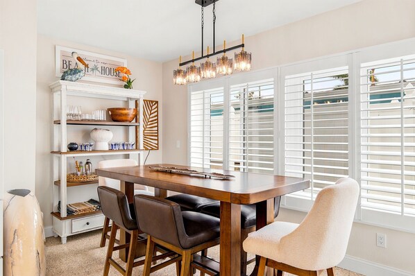 A dining area with a wooden table, chairs, a chandelier, and a window with blinds.
