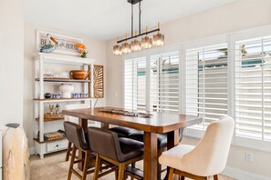 A dining area with a wooden table, chairs, a chandelier, and a window with blinds.