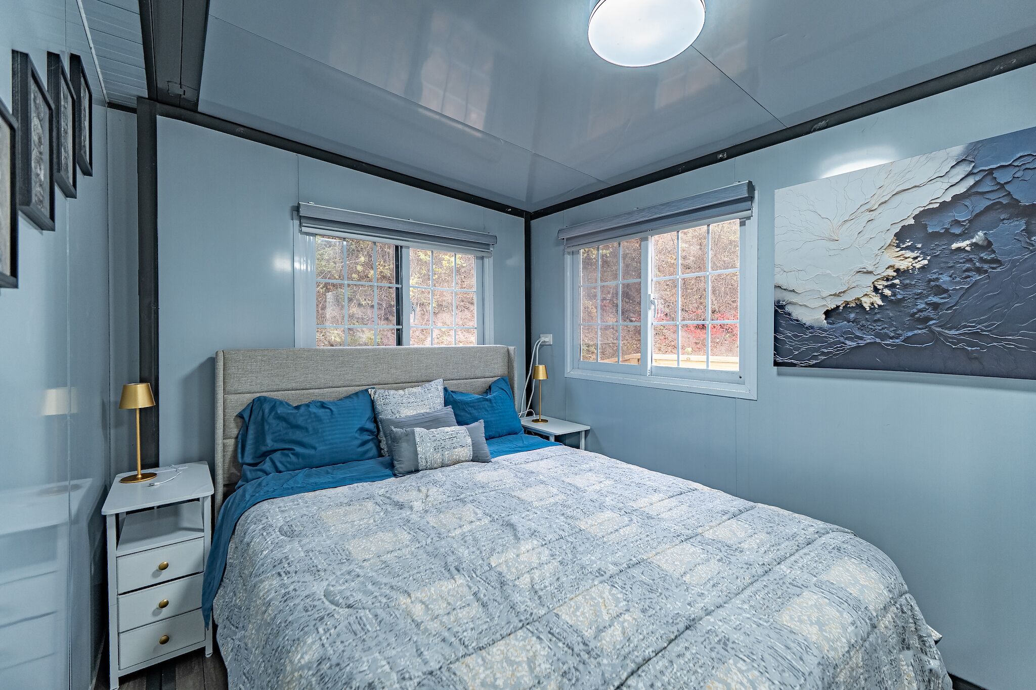 A bedroom with a bed, two windows, a nightstand, a lamp, and a wall-mounted artwork.