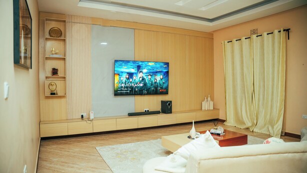 Una sala de estar moderna con una televisión de pantalla plana montada en la pared, un mueble de madera con objetos decorativos, un sofá blanco y un ventanal con cortinas.