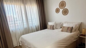 Een keurig opgemaakt bed met witte lakens, een houten vloer en een raam met dunne gordijnen.