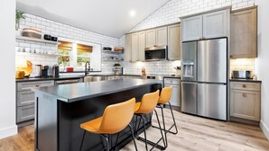 A modern kitchen with a central island, bar stools, stainless steel appliances, and white tiled backsplash.