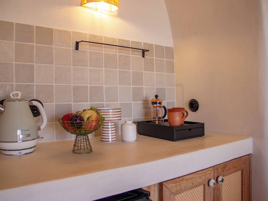 Une cuisine avec un mur recouvert de carreaux, une bouilloire, un panier de fruits, des tasses empilées et une machine à café.
