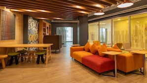 A modern lobby with a wooden ceiling, a central orange sofa, and a dining area with stools.