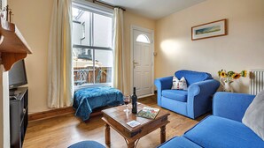 Een gezellige woonkamer met een houten salontafel, een blauwe bank en een blauwe fauteuil. Een televisie staat op een dressoir links, en er is een deur te zien op de achtergrond.