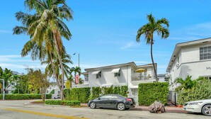 A street view with a white building, palm trees, and parked cars. - Chic 1-Bedroom Apt Steps from the Beach in Beautiful Miami Beach (Miami Beach)