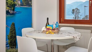 A dining area with a round table, white chairs, a clock on the wall, and a window with a view of a lake and mountains. - Residence Lilly-APT Fefe- Balcony lake view, Residence Lilly - APT Fefe - Balcone vista lago (Malcesine)