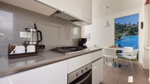 A modern kitchen with a gas stove, oven, and coffee maker. There is a dining area with a table and chairs, and a large window offering a scenic view of a lake and hills. - Residence Lilly-APT Fefe- Balcony lake view, Residence Lilly - APT Fefe - Balcone vista lago (Malcesine)