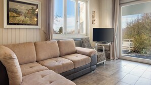 Ein Wohnzimmer mit einem beigefarbenen Sofa, einem Fernseher auf einem kleinen Tisch und einem großen Fenster mit Blick auf Bäume und ein Gebäude.