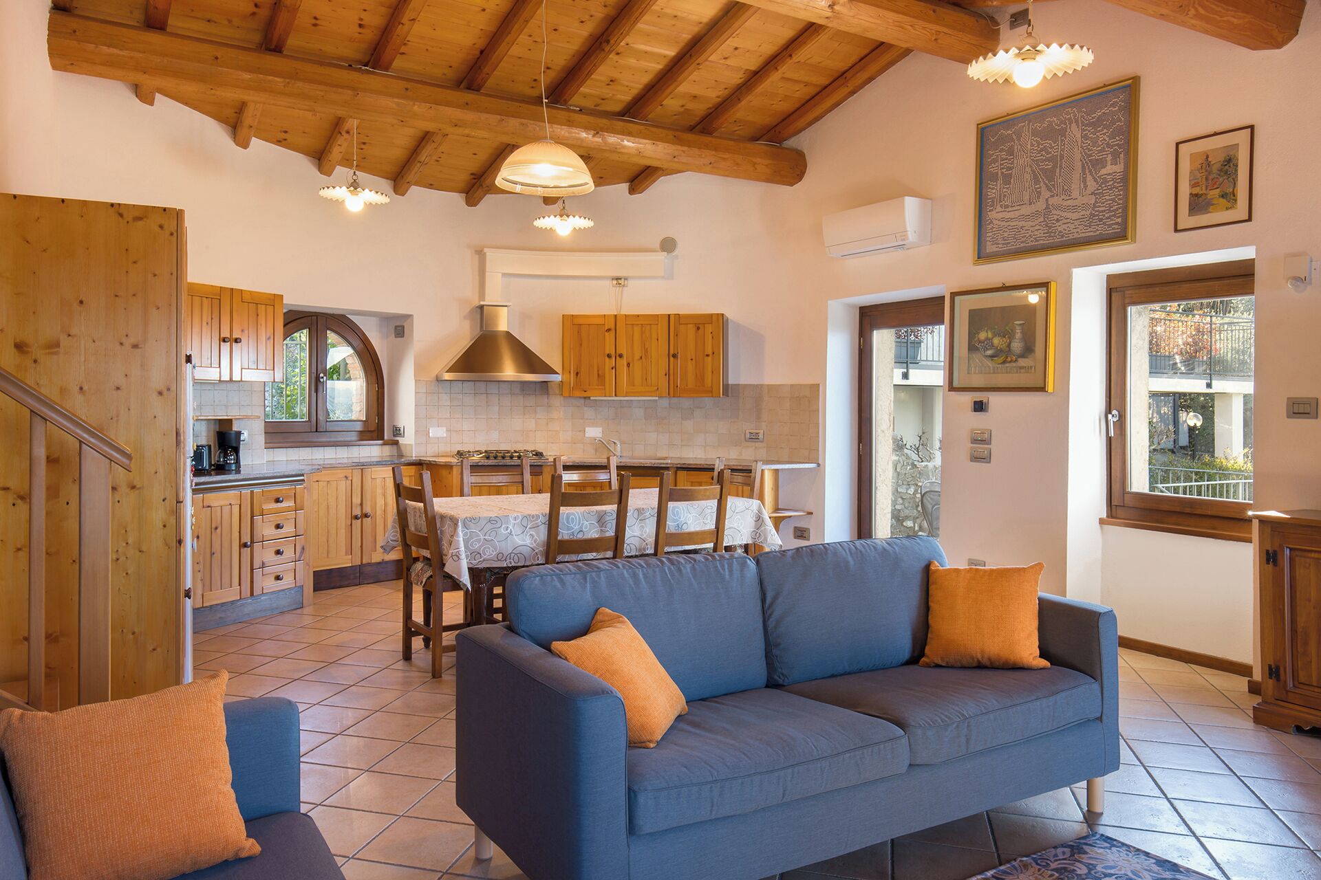 A cozy living room with a blue sofa and orange cushions, wooden cabinets, a tiled floor, and a kitchen area with wooden beams and pendant lights.