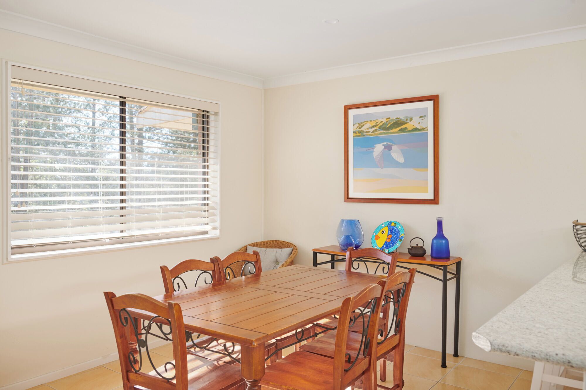 A dining area with a wooden table and chairs, a window with blinds, a framed painting, and decorative vases on a side table.