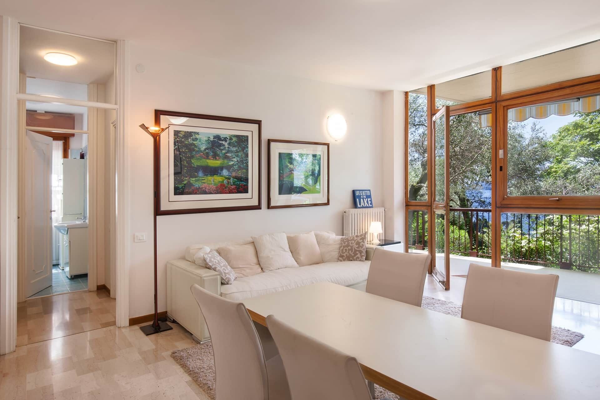 Un salon moderne avec un canapé blanc, une table à manger et des chaises. Deux tableaux encadrés ornent le mur, une fenêtre offre une vue sur la verdure et un radiateur est installé.