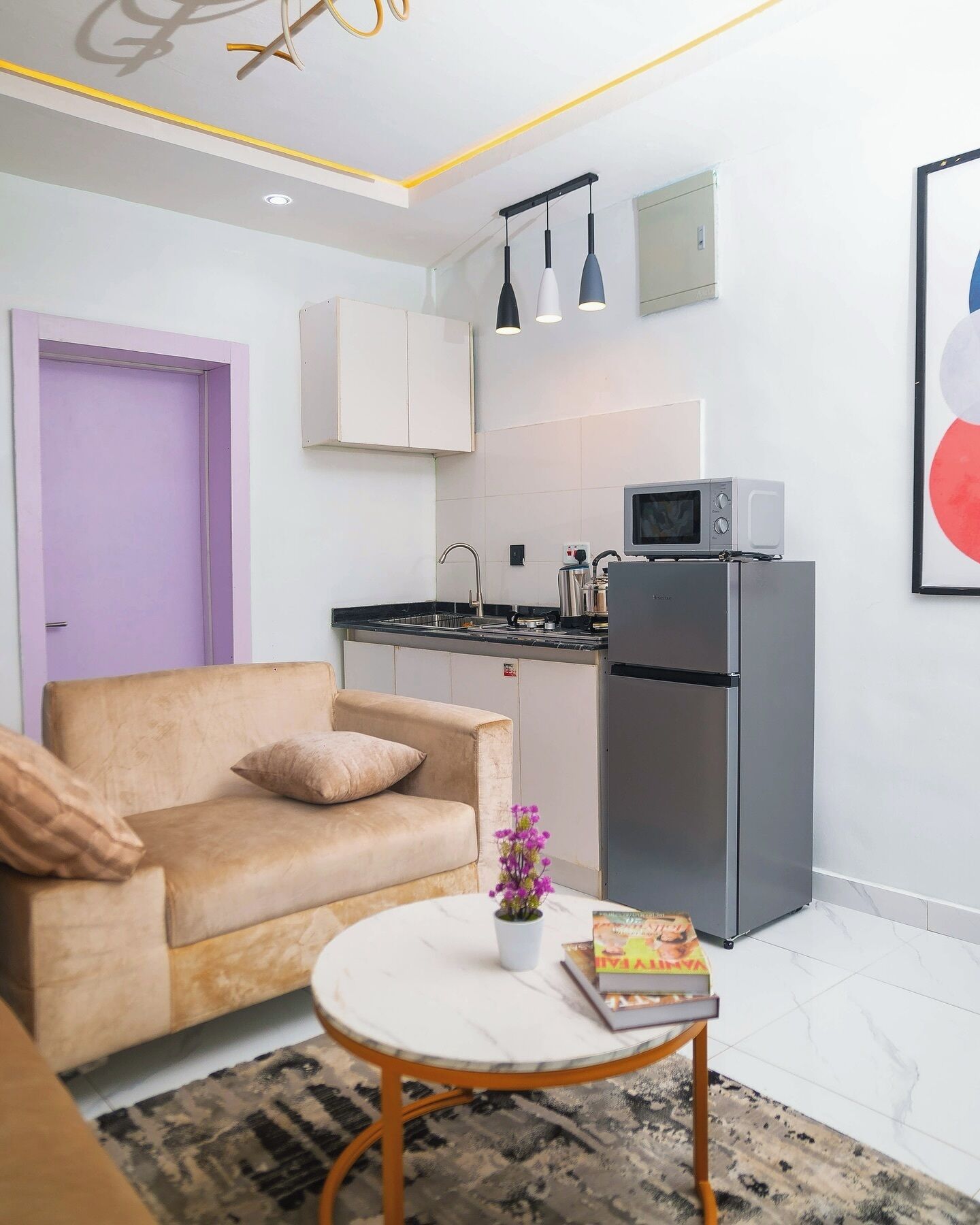 A modern living room with a beige sofa, a small round coffee table with a book and a flower, and a kitchen area with a refrigerator, microwave, and hanging pendant lights.