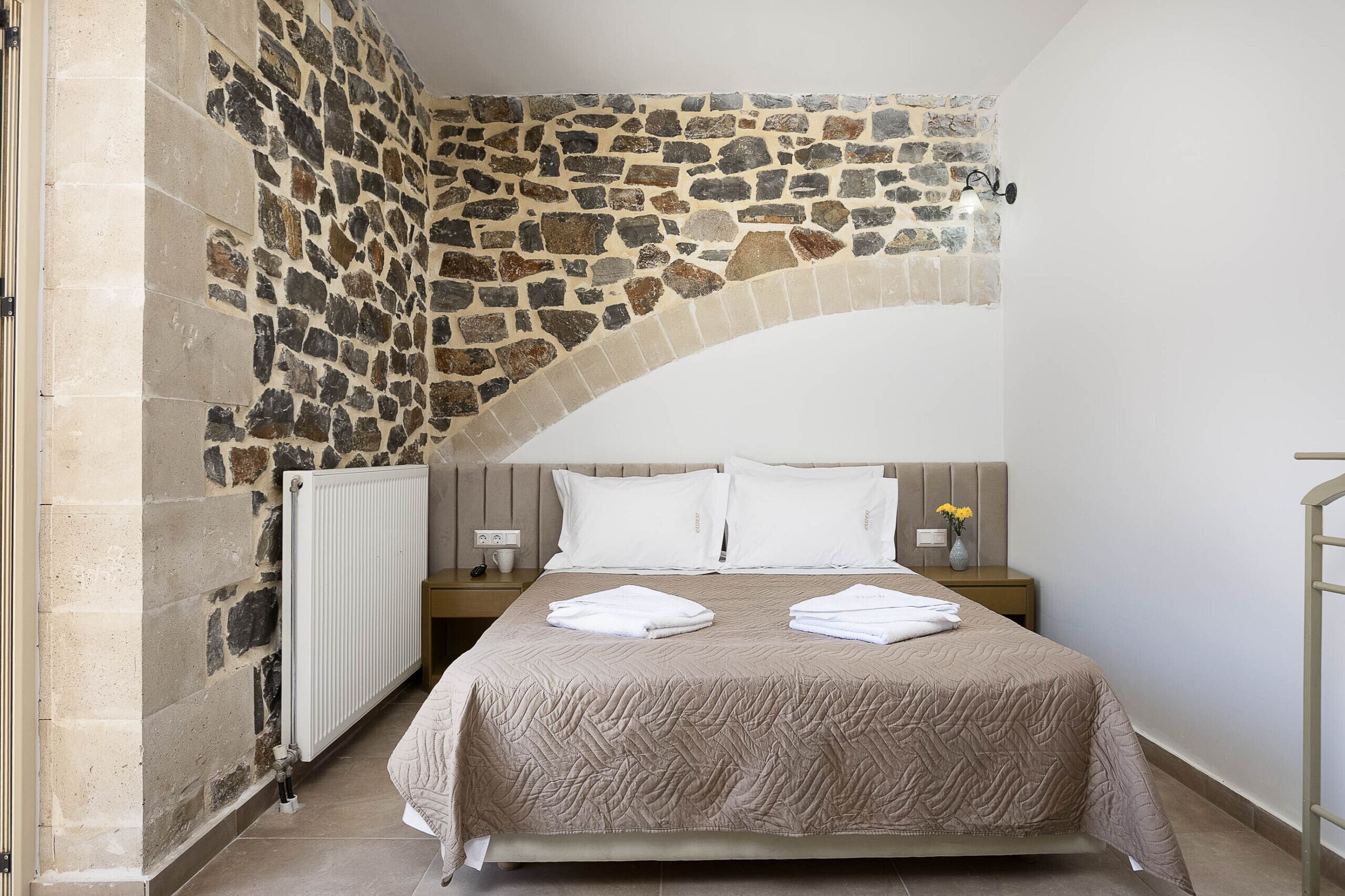 A bedroom with a stone wall, a bed with a beige quilt, two white pillows, a bedside table with a vase, and a nightstand with a lamp.
