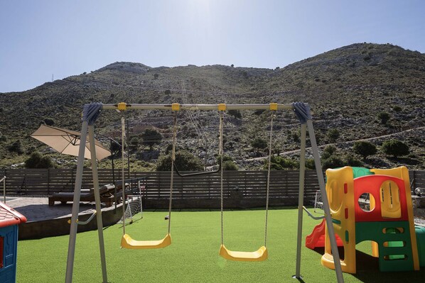 Un parque infantil con columpios y un tobogán, rodeado por una cerca de madera y una colina al fondo.