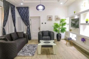 A modern living room with a dark brown sofa, a black armchair, a coffee table, and a patterned rug. There is a door with a decorative handle, a potted plant, and a wall-mounted TV. - Blue Velvet Apartments (Abuja)