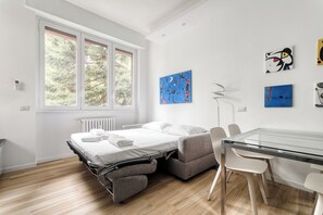 Un dormitorio moderno con una cama tipo sofá, una mesa de comedor de cristal y vistas a árboles a través de amplios ventanales.