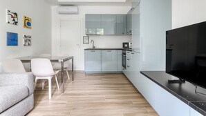 Una cocina moderna con gabinetes grises, una encimera negra y un televisor grande. Hay un comedor con una mesa y sillas blancas.