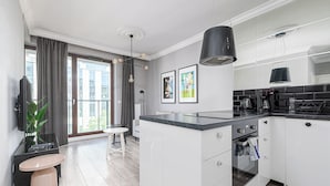 A modern kitchen with white cabinets, a black countertop, and a built-in oven. There is a dining area with a table and chairs, and a large window with curtains. - RentPlanet - Apartament Siedmiogrodzka (Warsaw)